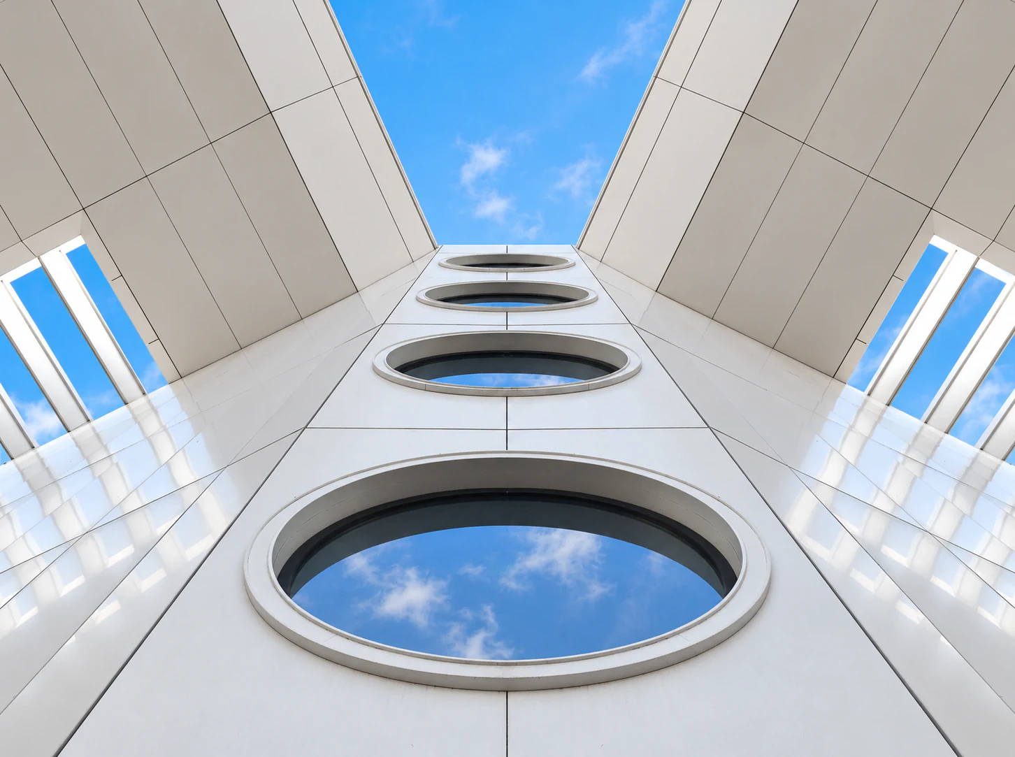 Upward view of circular glazing openings with cloud reflections, reflective cladding and projecting roof panels framing the blue sky
