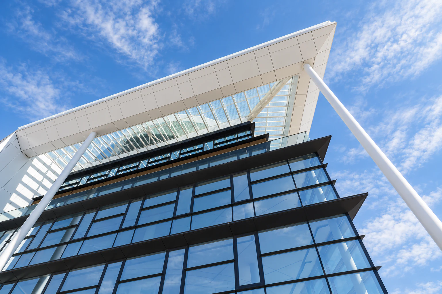 Upward view of glass façade with dark horizontal shading fins, slender white columns and projecting roof beneath a textured blue sky